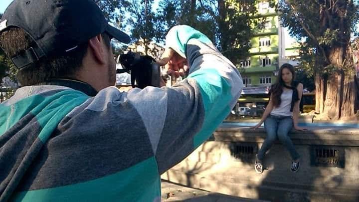 Berguim fotografando, de costas, uma modelo sentada em um muro de um parque.